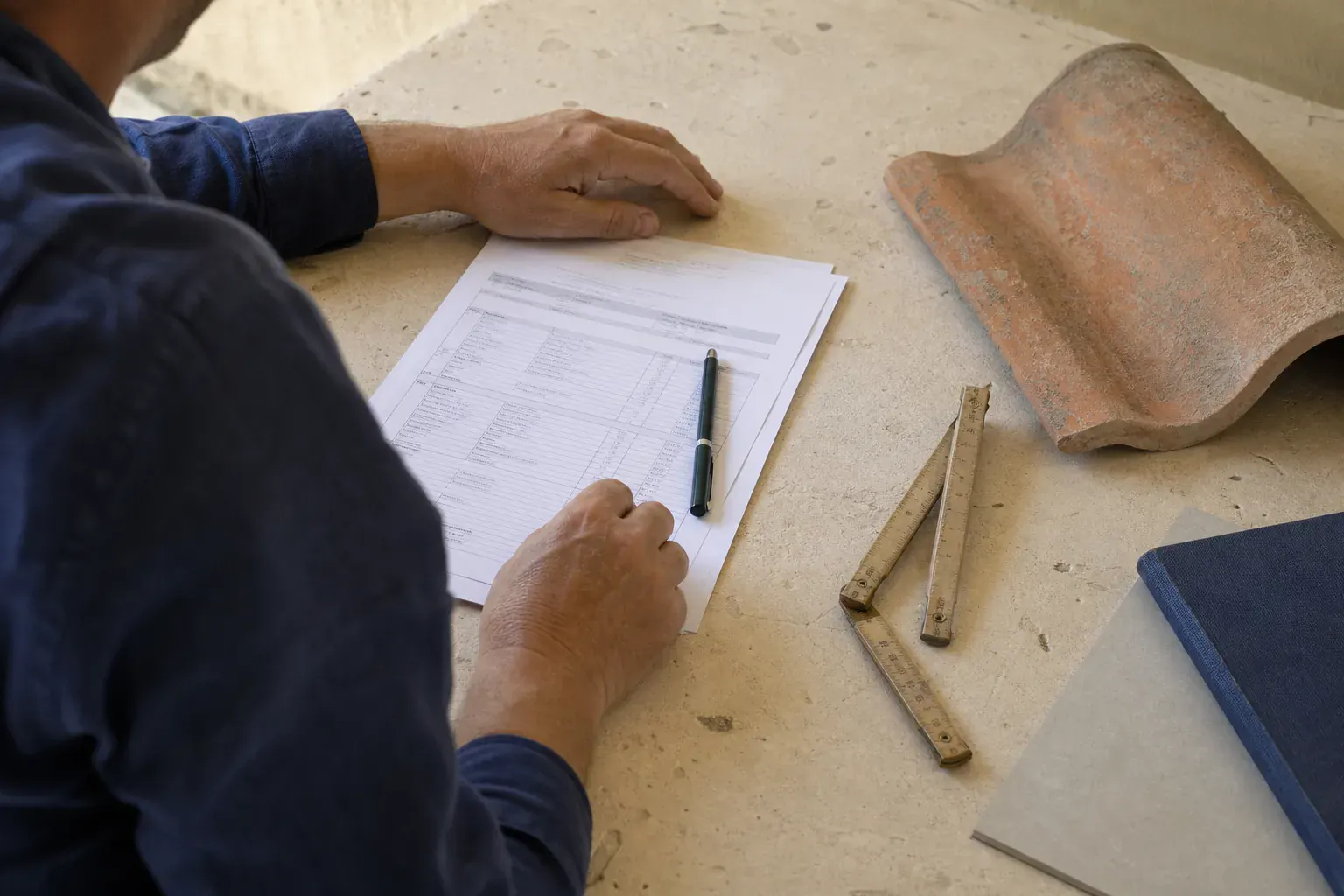 Devis détaillé poste par poste posé sur une table nue, stylo, mètre pliant et échantillon de tuile de terre cuite
